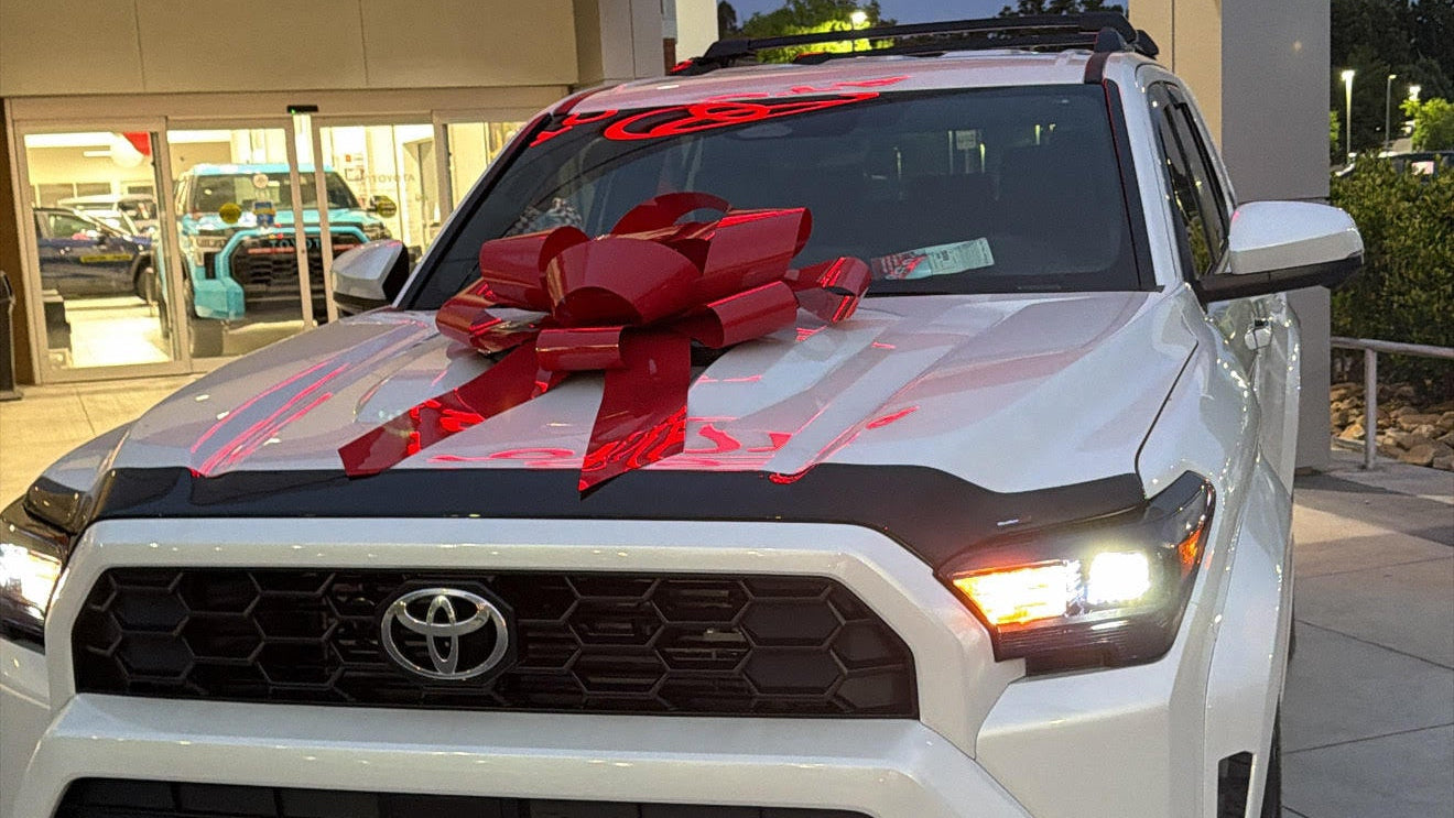 A white Toyota pickup truck parked outdoors in front of a dealership building, with a large red gift bow and red ribbon across the hood.
